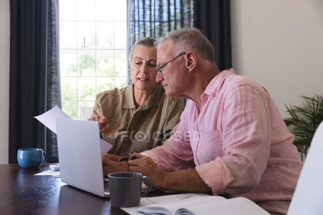 Casal idoso caucasiano na sala de estar sentado à mesa usando laptop, pagando contas e conversando. ficar em casa em isolamento durante o confinamento de quarentena. — Fotografia de Stock