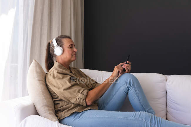 Mujer mayor caucásica feliz en la sala de estar sentado en el sofá con auriculares y el uso de teléfonos inteligentes. permanecer en casa aislado durante el bloqueo de cuarentena. - foto de stock