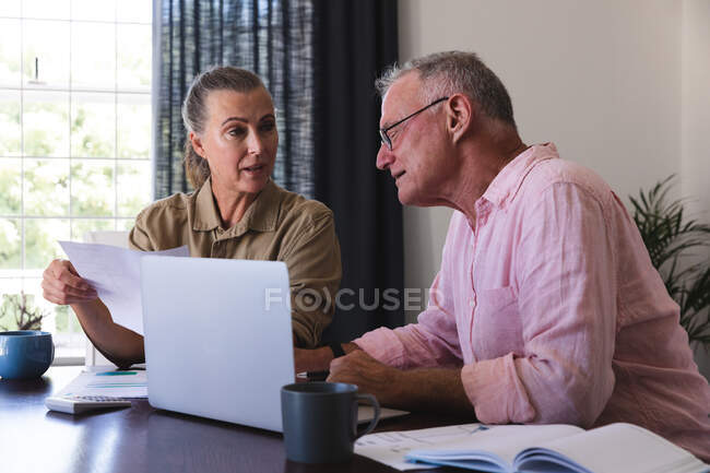 Kaukasisches Seniorenpaar im Wohnzimmer, das mit Laptop am Tisch sitzt, Rechnungen bezahlt und redet. Isolationshaft während der Quarantäne. — Stockfoto