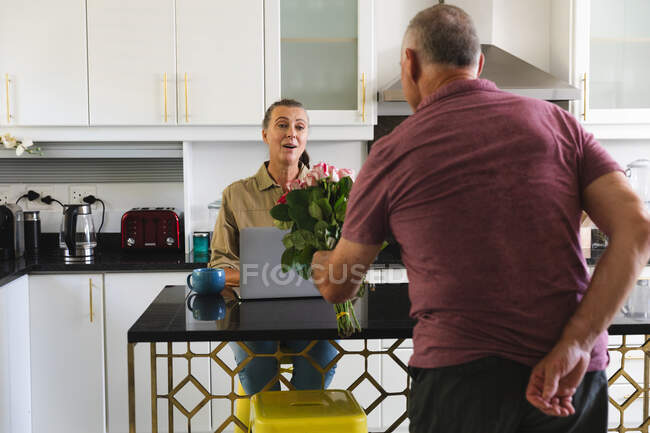 Счастливая белая старшая пара на кухне, женщина с ноутбуком, мужчина, дарящий ей букет цветов. оставаться дома в изоляции во время карантинной изоляции. — стоковое фото