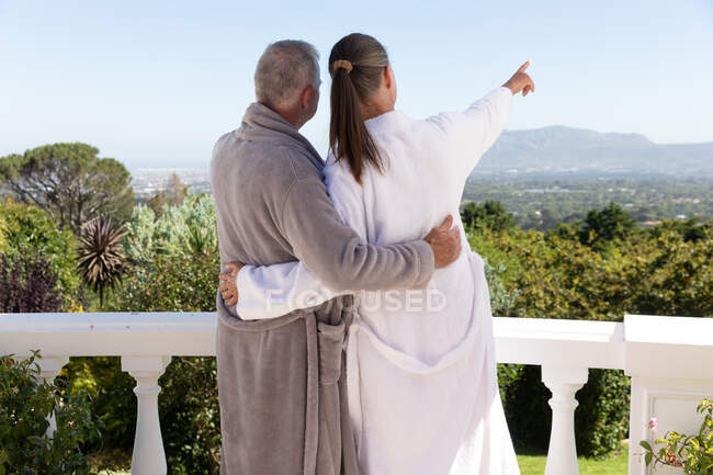Vista posteriore della coppia anziana caucasica che indossa accappatoi abbracciati sul balcone, donna che punta. stare a casa in isolamento durante la quarantena. — Foto stock