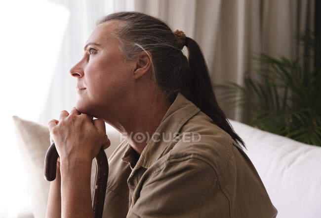 Caucasian senior woman in living room sitting on couch in thought, leaning chin on walking stick. staying at home in isolation during quarantine lockdown. — Stock Photo