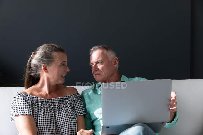 Счастливая старшая кавказская пара отдыхает в гостиной, сидя на диване с ноутбуком и разговаривая. оставаться дома в изоляции во время карантинной изоляции. — стоковое фото