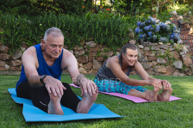 Heureux couple d'aînés caucasiens faisant de l'exercice dans le jardin, assis sur des nattes pratiquant le yoga. rester à la maison dans l'isolement pendant le confinement en quarantaine. — Photo de stock