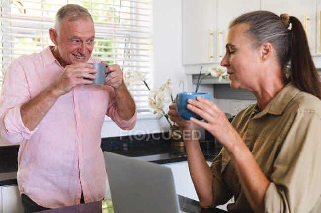 Feliz pareja de ancianos caucásicos en la cocina bebiendo café y hablando. permanecer en casa aislado durante el bloqueo de cuarentena. - foto de stock