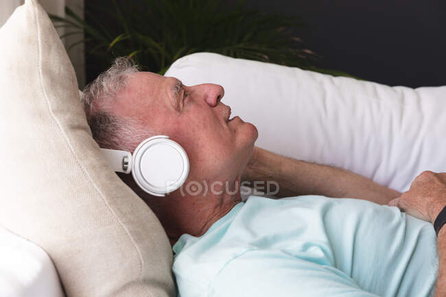 Heureux homme âgé caucasien dans le salon couché sur le canapé portant des écouteurs et souriant. rester à la maison dans l'isolement pendant le confinement en quarantaine. — Photo de stock