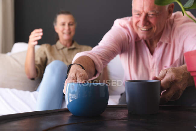 Щаслива кавказька старша пара розслабляється у вітальні, чоловік кладе чашки на стіл і посміхається. перебування вдома в ізоляції під час карантину . — стокове фото