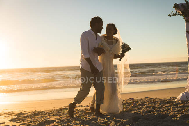 Coppia afroamericana innamorata che si sposa, che cammina sulla spiaggia tenendosi per mano al tramonto. amore, romanticismo e matrimonio in spiaggia vacanze estive. — Foto stock