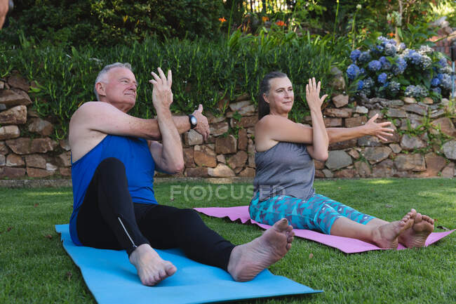 Casal sênior caucasiano feliz exercitando no jardim, sentado em tapetes praticando ioga. ficar em casa em isolamento durante o confinamento de quarentena. — Fotografia de Stock