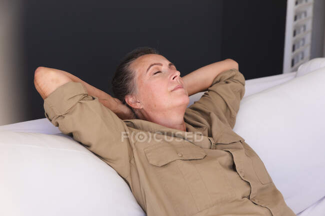 Mujer mayor caucásica feliz en la sala de estar tumbada en el sofá con los brazos detrás de la cabeza y los ojos cerrados. permanecer en casa aislado durante el bloqueo de cuarentena. - foto de stock