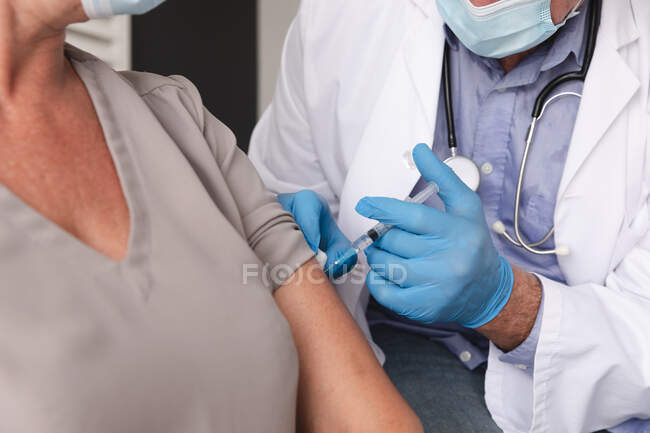 Midsection of caucasian male doctor using face mask giving female patient covid 19 vaccination. profesional médico en el trabajo durante la pandemia del coronavirus covid 19. - foto de stock