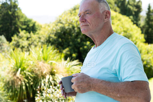 Homem idoso caucasiano em pé no jardim ensolarado, segurando xícara de café e olhando para longe. ficar em casa em isolamento durante o confinamento de quarentena. — Fotografia de Stock