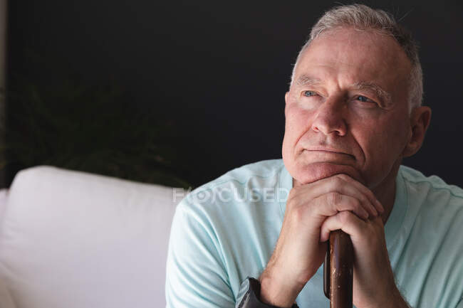 Caucasian senior man in living room sitting on couch in thought, leaning chin on walking stick. staying at home in isolation during quarantine lockdown. — Stock Photo