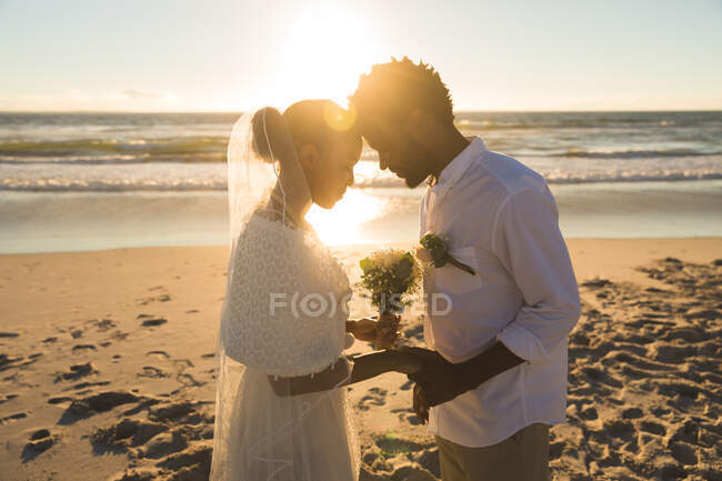 Happy african american couple in love getting married, touching foreheads on beach during sunset. love, romance and beach break summer holiday. — Stock Photo