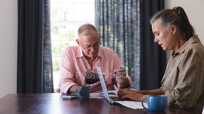 Casal idoso caucasiano na sala de estar sentado à mesa de trabalho, usando laptop e tablet. ficar em casa em isolamento durante o confinamento de quarentena. — Fotografia de Stock