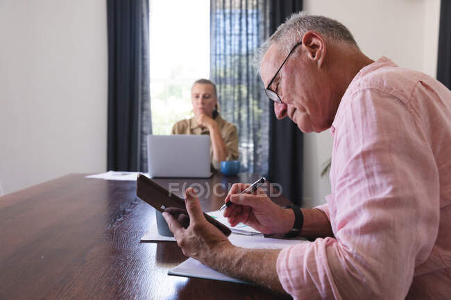 Casal idoso caucasiano na sala de estar sentado à mesa de trabalho, usando laptop e tablet. ficar em casa em isolamento durante o confinamento de quarentena. — Fotografia de Stock