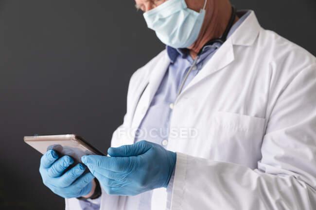 Midsection of caucasian senior male doctor using face mask and surgical gloves using tablet. profesional médico en el trabajo durante la pandemia del coronavirus covid 19. - foto de stock