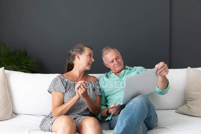 Feliz pareja mayor caucásica sentada en la sala de estar mirando a la computadora portátil hablando y sonriendo. permanecer en casa aislado durante el bloqueo de cuarentena. - foto de stock