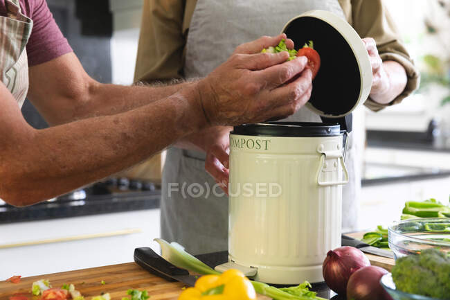 Die Mitte des kaukasischen Seniorehepaares in der Küche trägt Schürzen, die organischen Abfall in die Komposttonne werfen. Isolationshaft während der Quarantäne. — Stockfoto