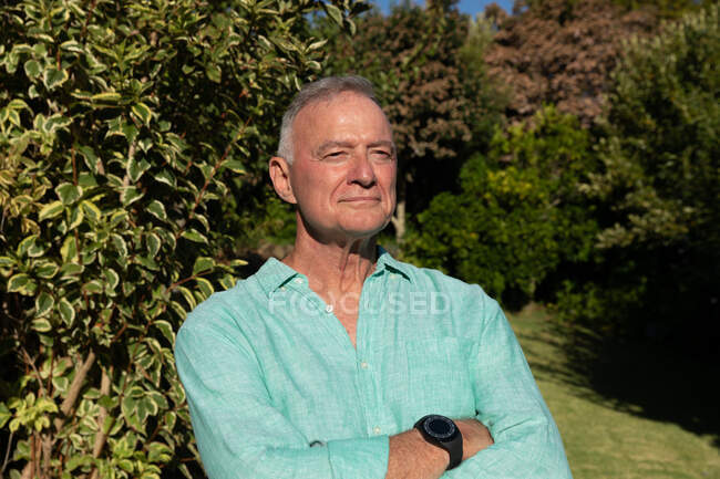 Glücklicher kaukasischer Senior, der mit verschränkten Armen lächelnd im sonnigen Garten steht. Isolationshaft während der Quarantäne. — Stockfoto