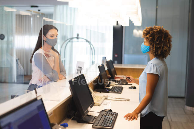 Diversas empresarias con máscaras en la cara hablando con la recepcionista en el hotel. viaje de negocios hotel durante coronavirus covid 19 pandemia. - foto de stock