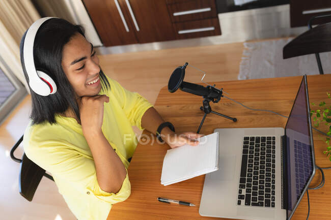 Femme transgenre mixte portant des écouteurs faisant podcast à l'aide d'un ordinateur portable, souriant, tenant des notes. rester à la maison dans l'isolement pendant le confinement en quarantaine. — Photo de stock