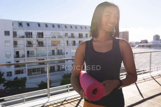 Gemischte Rasse Transgender-Frau praktiziert Yoga lächelnd auf Dachterrasse in der Sonne hält Matte. Isolationshaft während der Quarantäne. — Stockfoto