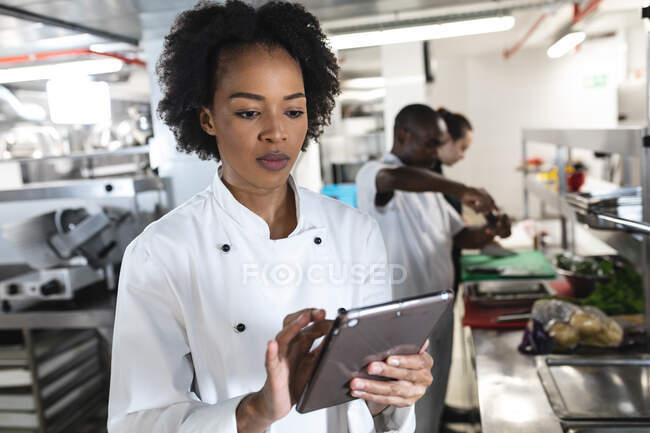 Gemischte Rasse Profi-Koch Blick auf Tablet mit Kollegen im Hintergrund. Arbeit in einer belebten Restaurantküche. — Stockfoto
