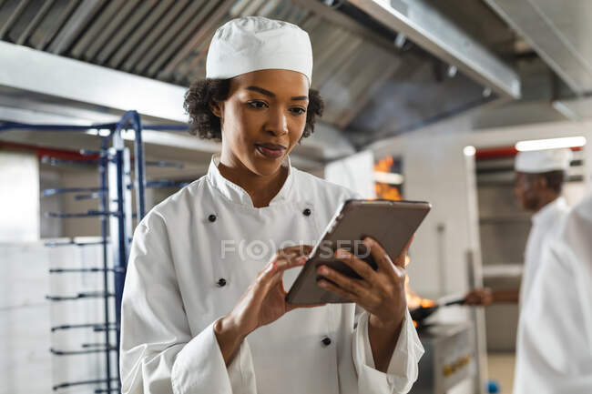 Портрет женщины-повара смешанной расы, использующей планшет с коллегой в фоновом режиме. работа на кухне ресторана. — стоковое фото