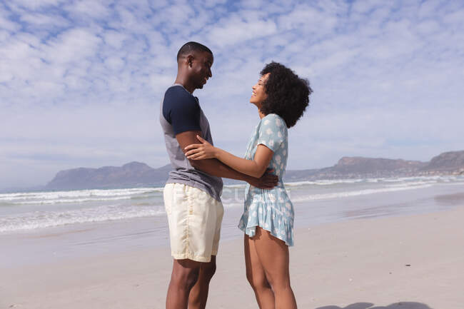 African american couple standing and looking at each other at the beach smiling. healthy outdoor leisure time by the sea. — Fotografia de Stock