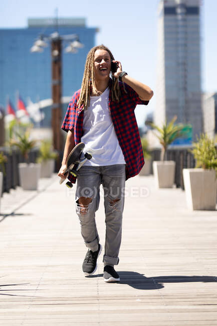 Buon uomo razza mista con dreadlocks portando skateboard parlando su smartphone in strada. nomade digitale, in giro per la città. — Foto stock