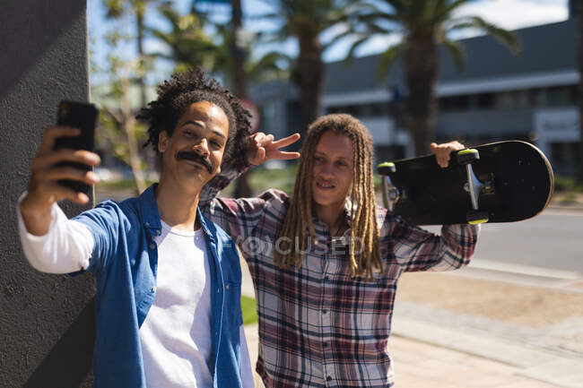 Two happy mixed race male friends holding skateboard in the street and taking selfie. digital nomad, out and about in the city. — Stock Photo