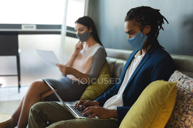 Diverse male and female of business colleagues wearing face masks sitting on sofa working. casual meeting in business lounge during coronavirus covid 19 pandemic. — Stock Photo