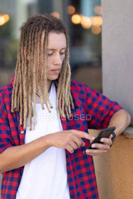 Masculino de raza mixta con rastas usando smartphone en la calle. nómada digital, fuera y alrededor de la ciudad. - foto de stock