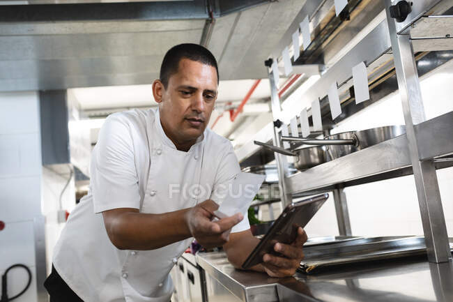 Chef profesional de carrera mixta mirando orden y tableta. trabajando en una cocina ajetreada. - foto de stock