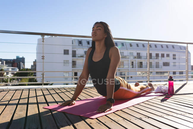 Женщина-трансгендер смешанной расы практикует йогу, растягиваясь на террасе на крыше на солнце. оставаться дома в изоляции во время карантинной изоляции. — стоковое фото