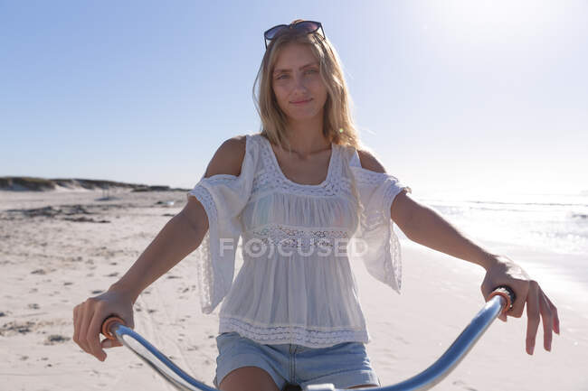 Lächelnde kaukasische Frau mit Sonnenbrille, weißem Oberteil und kurzen Hosen, die auf einem Fahrrad am Strand fährt. gesunde Freizeit im Freien am Meer. — Stockfoto