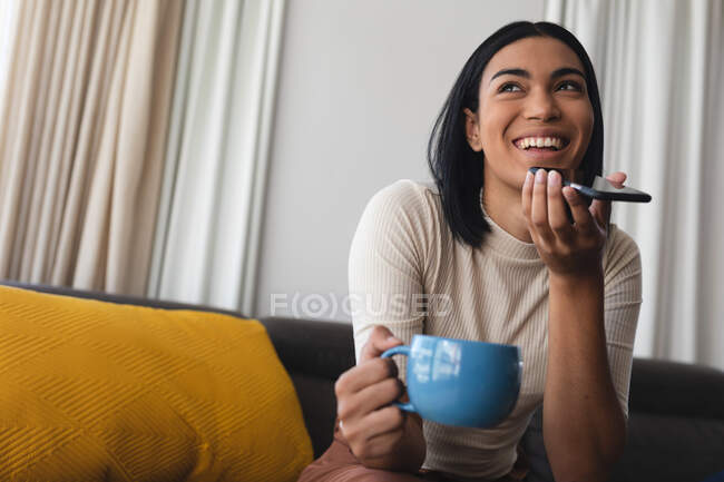 Glückliche Transgender-Frau mit gemischter Rasse, die es sich im Wohnzimmer gemütlich macht und auf der Couch sitzt und telefoniert. Isolationshaft während der Quarantäne. — Stockfoto