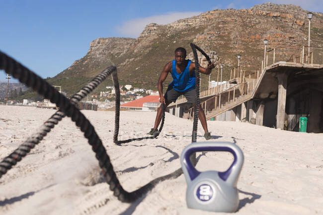 African american man exercising with rope and kettle on beach. healthy outdoor lifestyle fitness training. — Foto stock