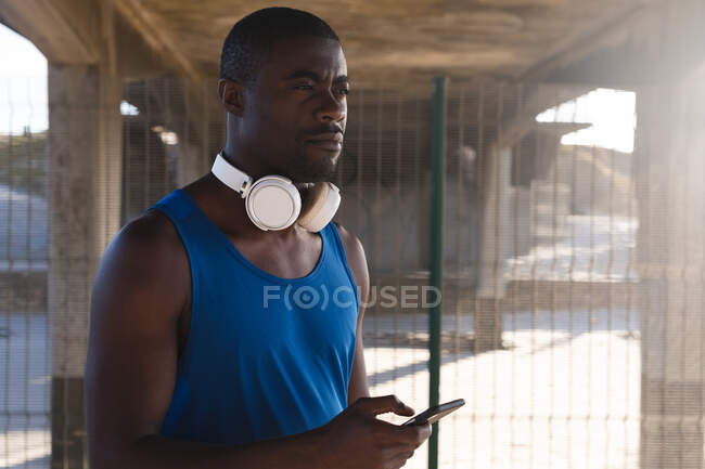Hombre afroamericano haciendo ejercicio al aire libre, usando auriculares, usando smartphone bajo puente. estilo de vida al aire libre saludable entrenamiento fitness. - foto de stock