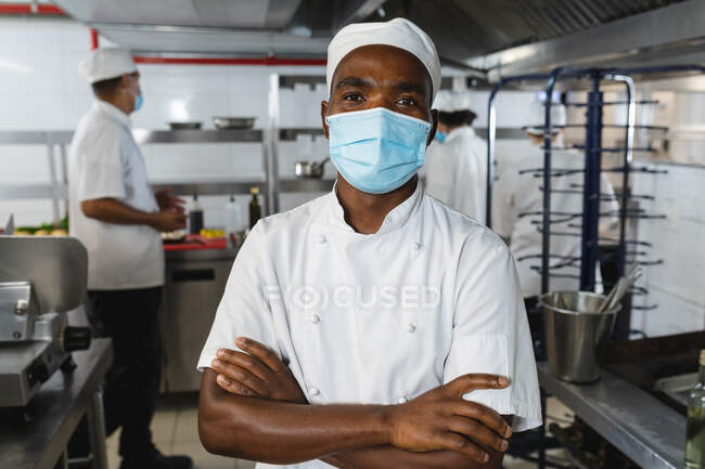 Retrato de un chef profesional afroamericano que usa mascarilla facial con colegas de fondo. trabajando en una ajetreada cocina de restaurante durante coronavirus covid 19 pandemia. - foto de stock