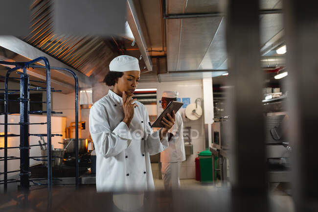 Портрет женщины-повара смешанной расы, использующей планшет с коллегой в фоновом режиме. работа на кухне ресторана. — стоковое фото