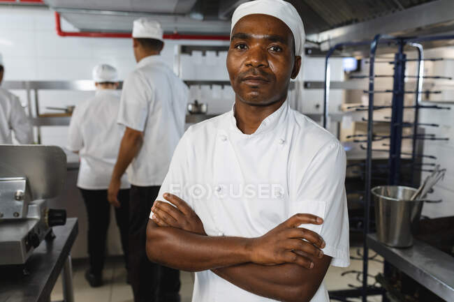 Porträt eines afrikanisch-amerikanischen Profikochs mit Kollegen im Hintergrund. Arbeit in einer belebten Restaurantküche. — Stockfoto