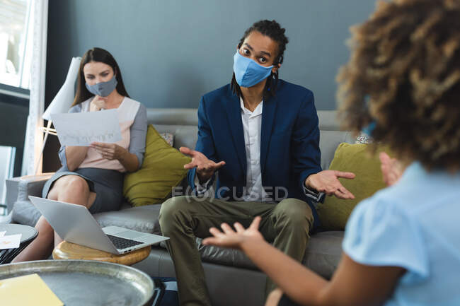 Diverse group of business colleagues wearing face masks sitting on sofa working. casual meeting in business lounge during coronavirus covid 19 pandemic. — Stock Photo