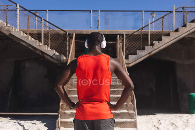 Visão traseira do homem afro-americano se exercitando, usando fones de ouvido na praia. treinamento de fitness ao ar livre saudável. — Fotografia de Stock