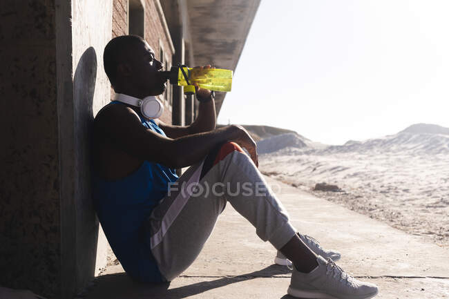 Afroamerikanischer Mann, der im Freien trainiert, Kopfhörer trägt, sich ausruht, Wasser trinkt. gesundes Outdoor-Fitness-Training. — Stockfoto