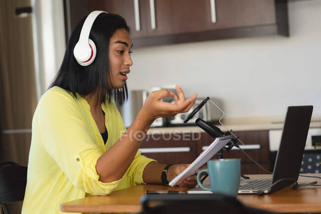 Mulher transgênero de raça mista usando fones de ouvido fazendo podcast usando laptop, falando, segurando notas. ficar em casa em isolamento durante o confinamento de quarentena. — Fotografia de Stock