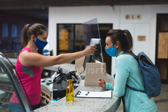 Female receptionist and customer wearing masks checking temperature over the counter at gym. fitness and leisure time at gym during coronavirus covid 19 pandemic. — Stock Photo