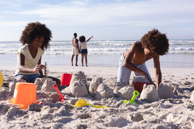 Afrikanisch-amerikanische Kinder beim Spielen mit Sand am Strand. mit den Eltern im Hintergrund. Familienfreizeit im Freien am Meer. — Stockfoto