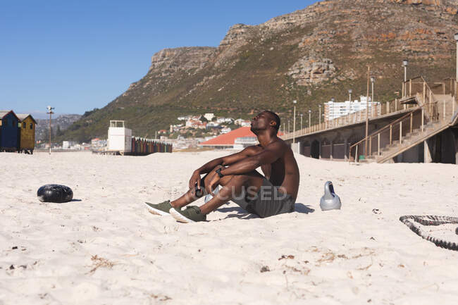 African american man exercising, resting on beach on sunny day. healthy outdoor lifestyle fitness training. — Stock Photo